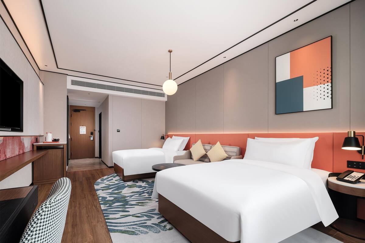 A modern hotel room with a large bed, a smaller bed, a desk with a TV, and a bathroom visible through an open door.