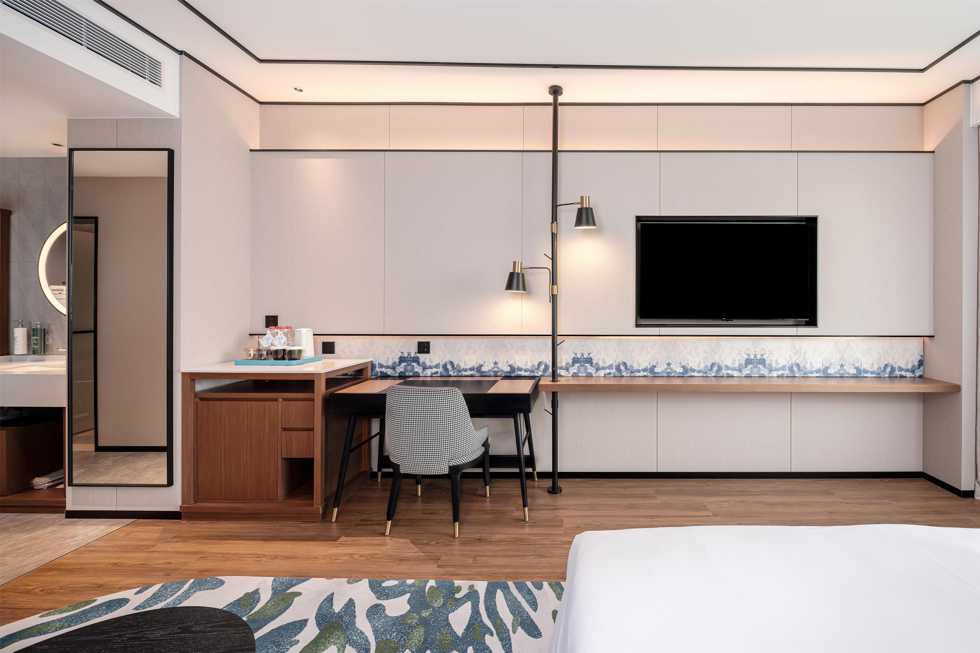 A modern hotel room with a wooden desk, a chair, a flat-screen TV mounted on the wall, and a bed with a white bedspread.