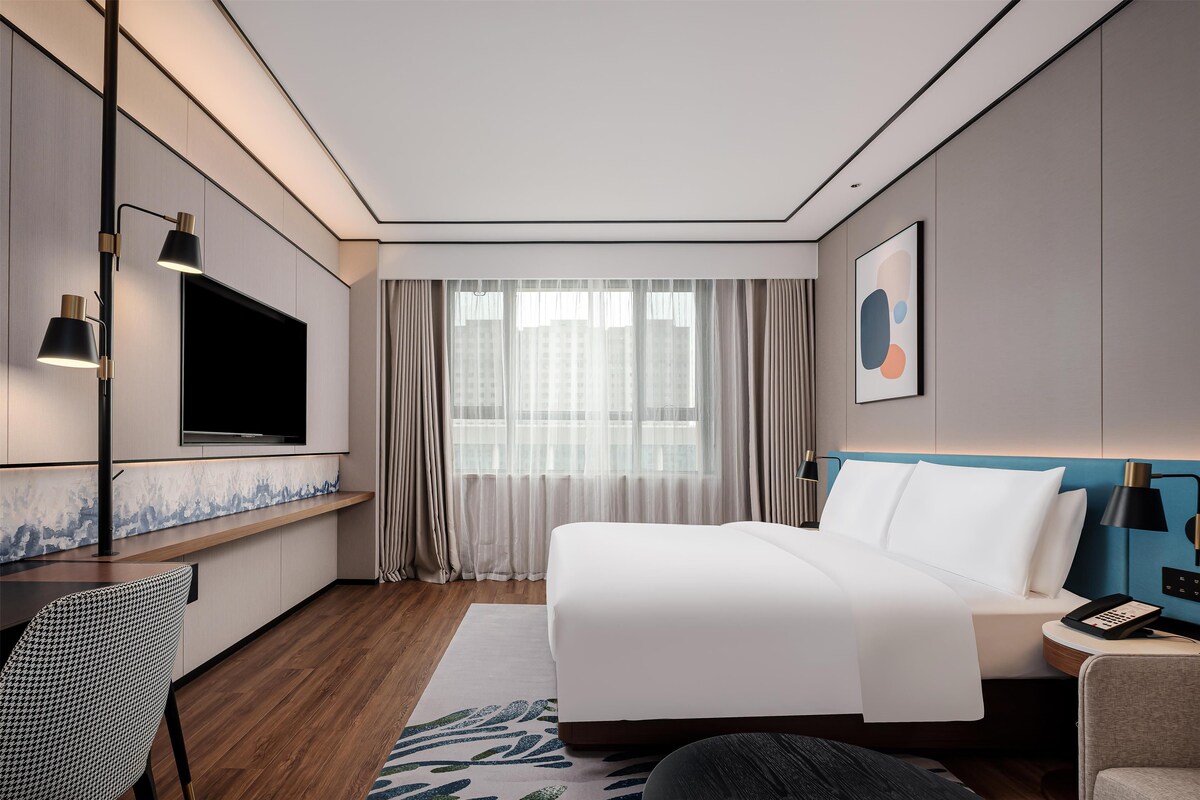 Habitación de hotel moderna con una cama grande, un televisor de pantalla plana montado en la pared, un escritorio con silla y una ventana con cortinas.