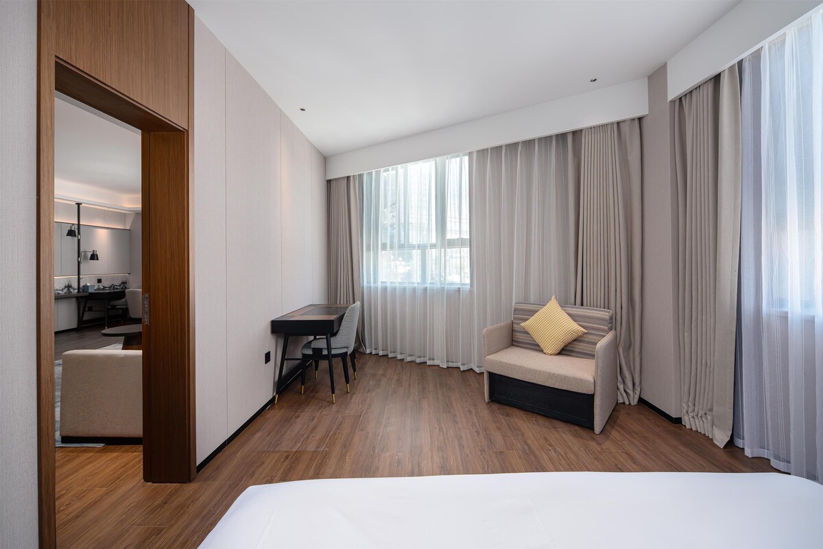 A hotel room with a bed, a desk, a chair, a sofa, and a large window with curtains.