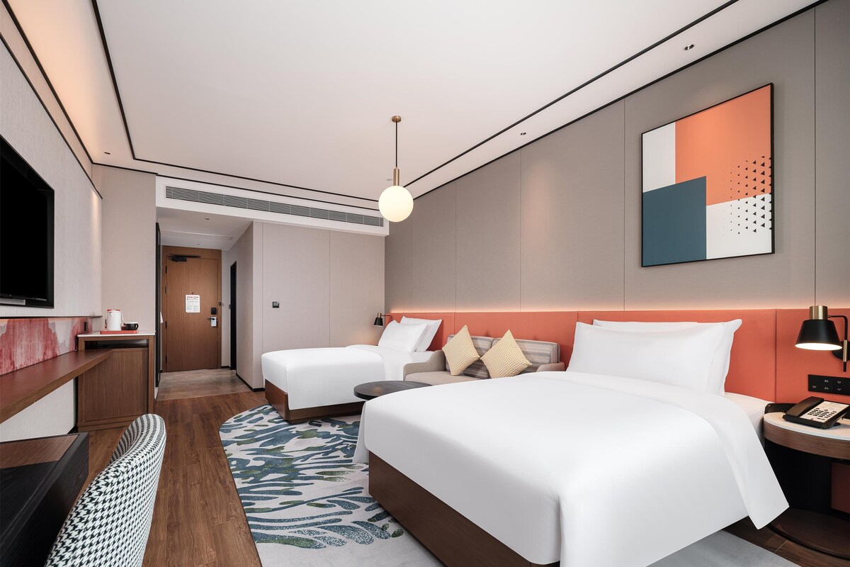 A modern hotel room with a large bed, a smaller bed, a desk with a TV, and a bathroom visible through an open door.