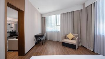 Quarto de hotel com cama, escrivaninha, cadeira, sofá e uma janela grande com cortinas.