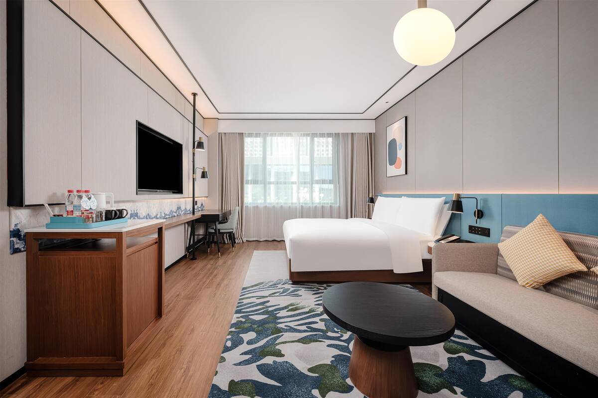 A modern hotel room with a bed, sofa, dining table, and TV.