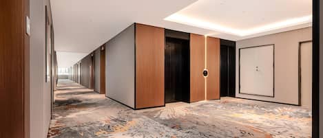A modern hotel hallway with wooden paneling, a patterned carpet, and a central elevator.