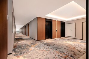A modern hotel hallway with wooden paneling, a patterned carpet, and a central elevator. - Hilton Garden Inn Yanqi Downtown (Hejing)
