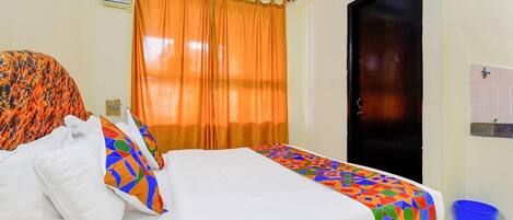 A hotel room with a bed, orange curtains, a black door, and a blue trash bin.