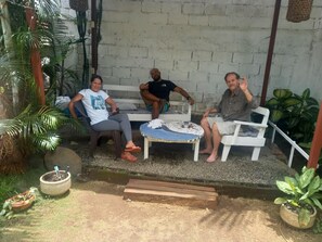 Three people seated on white plastic chairs in a covered outdoor area with a concrete wall and potted plants. - Hotel Cortez (San José)