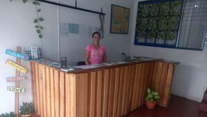 A reception desk with a wooden finish, a woman standing behind it, and various signs including 'Costa Rica', 'Welcome', 'Pura Vida', and 'Love'. - Hotel Cortez (San José)