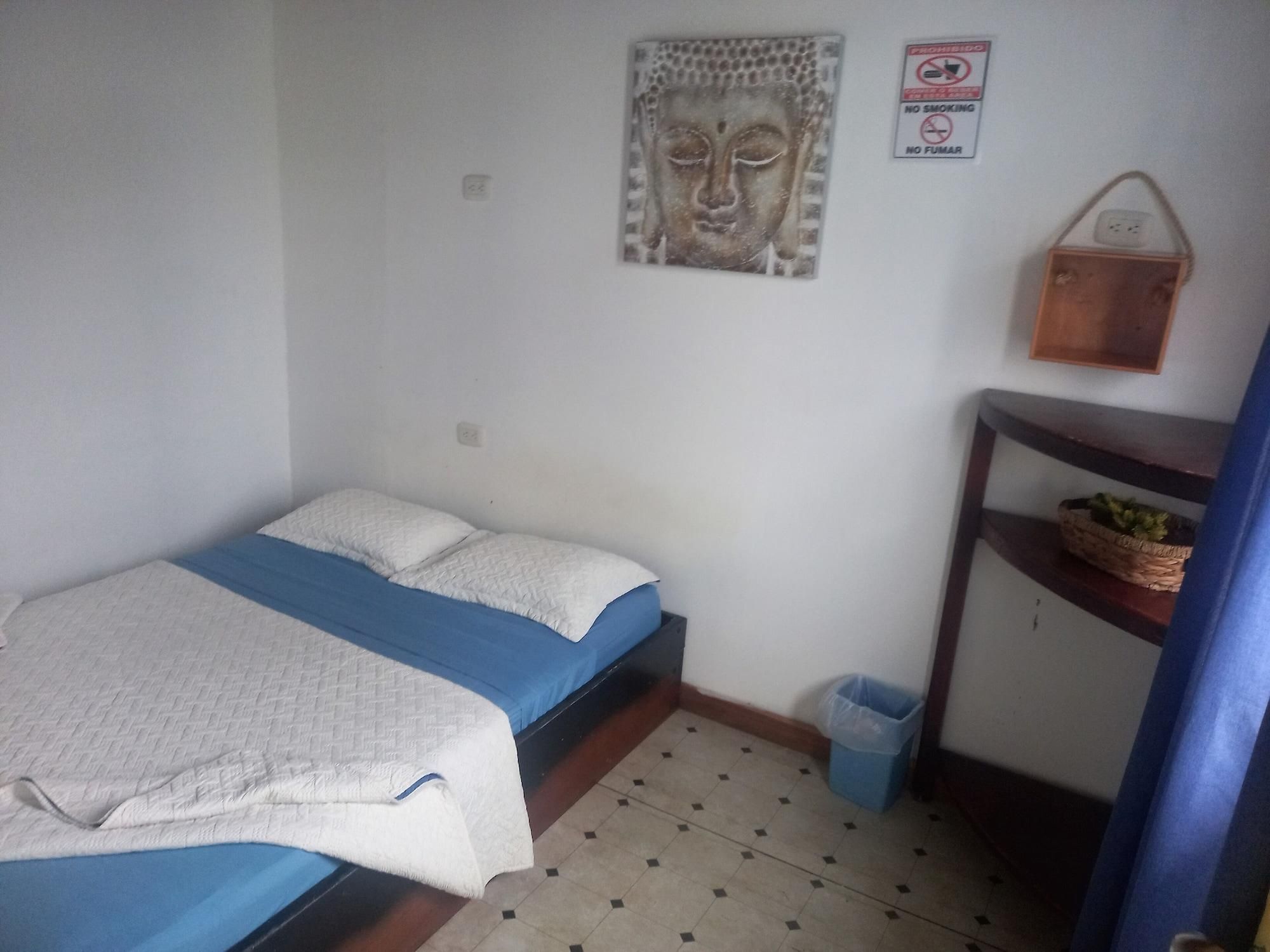 Una habitación pequeña y sencilla con una cama, una estatua de Buda en la pared, un cartel de prohibido fumar y una pequeña estantería de madera con una cesta.