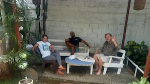 Three people seated on white plastic chairs in a covered outdoor area with a concrete wall and potted plants. - Hotel Cortez (San Jos é)