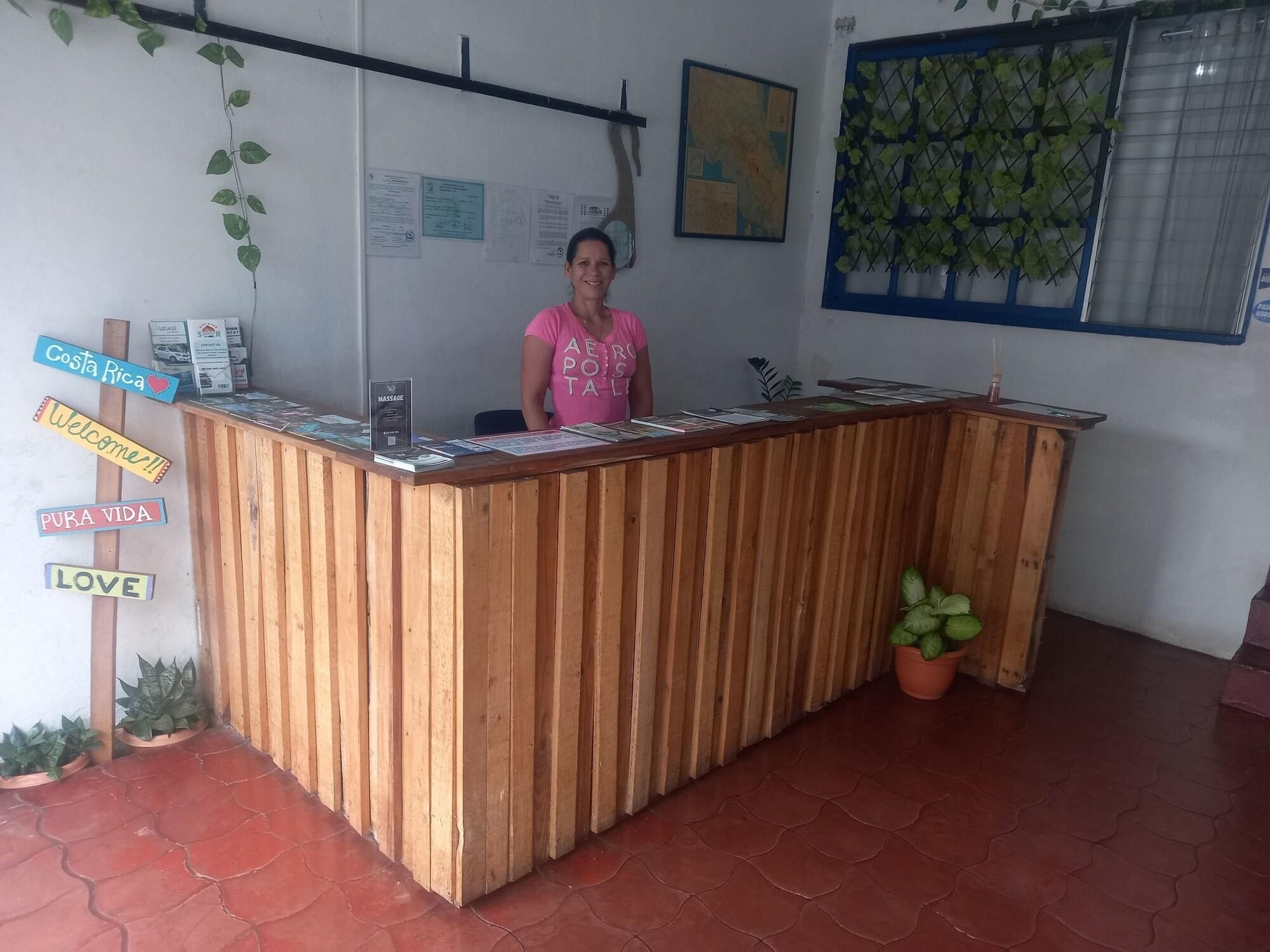 A reception desk with a wooden finish, a woman standing behind it, and various signs including 'Costa Rica', 'Welcome', 'Pura Vida', and 'Love'.