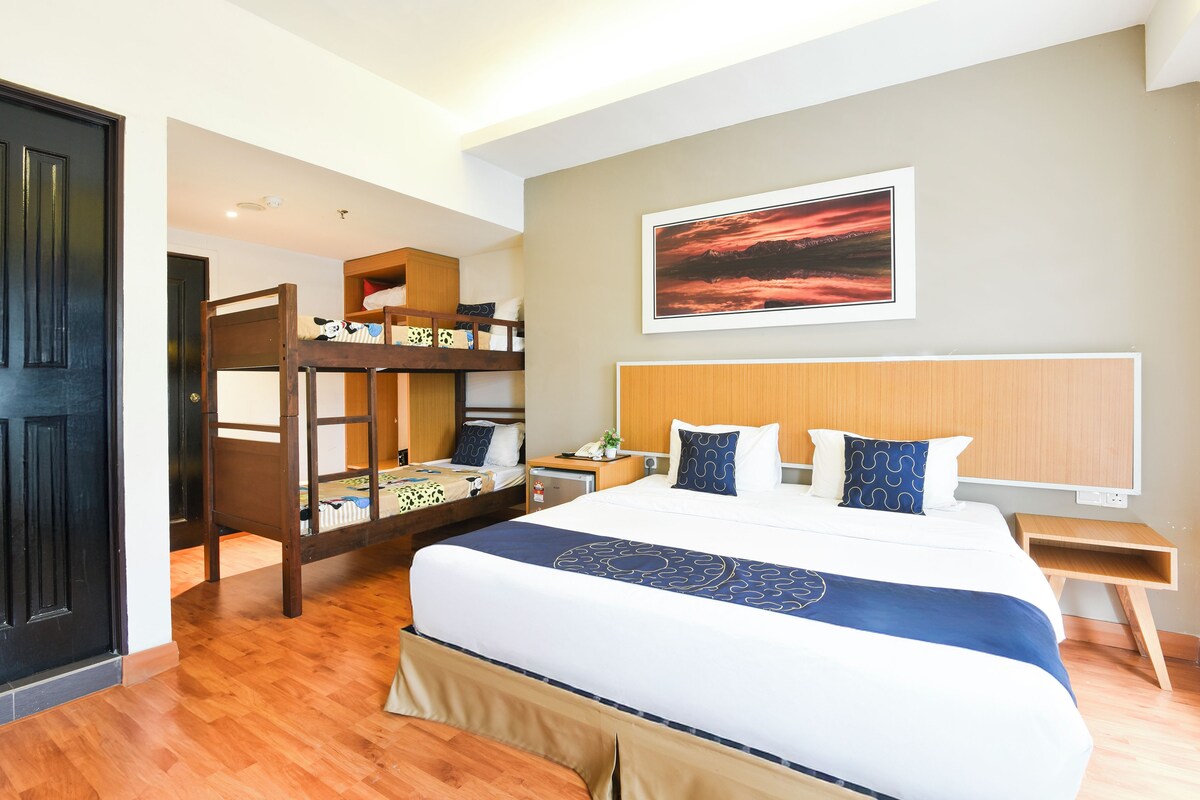 A hotel room with a bunk bed, a large bed with a blue and white bedspread, a wooden headboard, a bedside table with a lamp, and a framed picture on the wall.