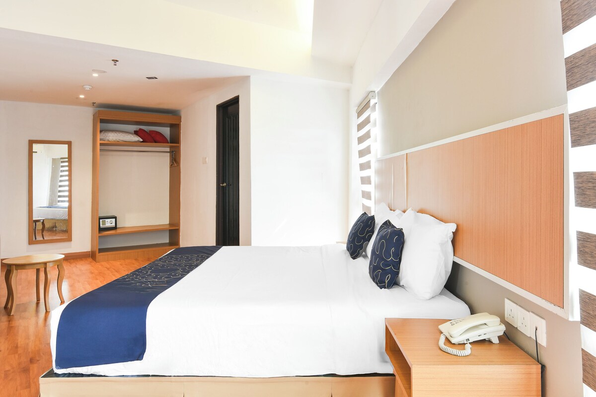 A hotel room with a large bed, a bedside table with a phone, a wardrobe, and a small table.