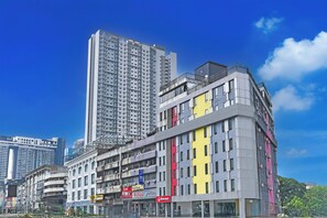 Un edificio moderno de varios pisos con espacios comerciales y residenciales, que destaca por una llamativa banda amarilla vertical y un balcón grande en los pisos superiores.