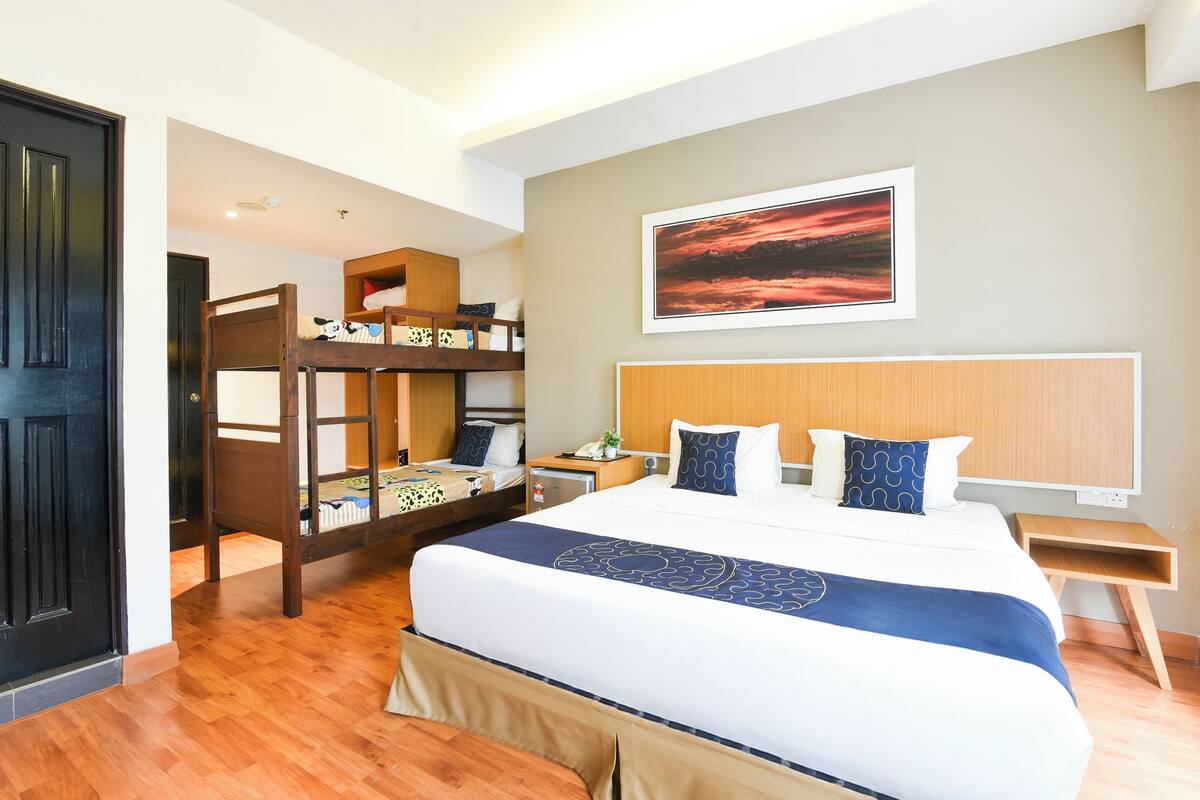 Een hotelkamer met een stapelbed, een groot bed met een blauw-witte sprei, een houten hoofdbord, een nachtkastje met een lamp en een ingelijst beeld aan de muur.