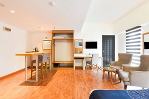 Una habitación de hotel moderna con un escritorio de madera, una cama, una zona de estar con dos sillones, una mesita y un televisor montado en la pared.