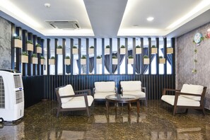 Un hall d’entrée avec un sol en marbre, un espace détente avec des fauteuils et une table, un mur avec des stores verticaux et une horloge.