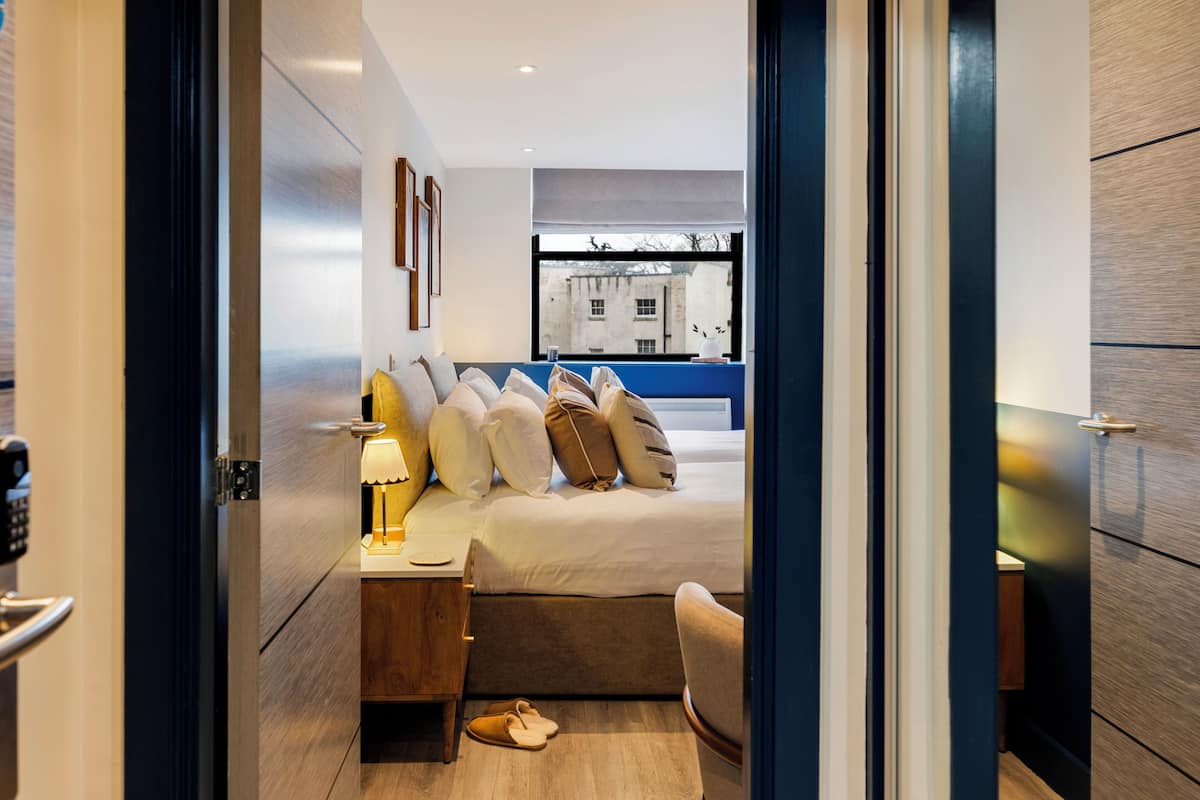 Una habitación de hotel moderna con una cama bien hecha, una mesita de noche con lámpara, una silla y vistas a la ciudad a través de un gran ventanal.