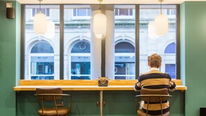 A person seated at a bar with a view of a building through large windows. - Clifton Village - Your Apartment (Bristol)