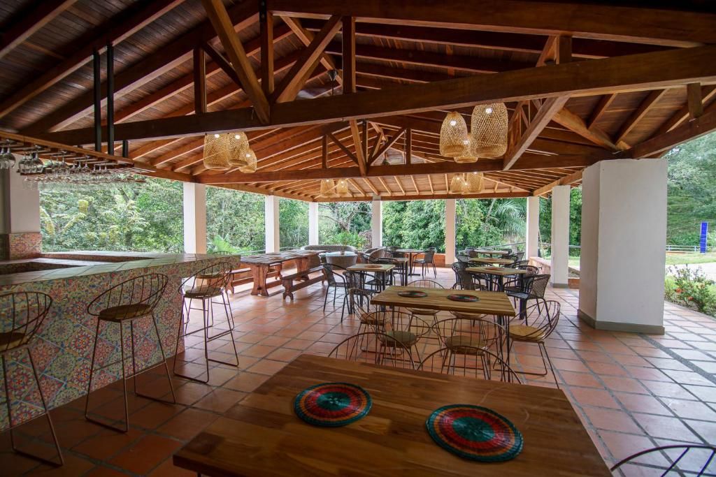 Un área de comedor al aire libre con mesas y sillas de madera, una barra con taburetes y grandes ventanales que ofrecen vistas a zonas verdes.
