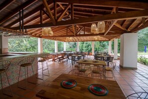 An open-air dining area with wooden tables and chairs, a bar with stools, and large windows offering a view of greenery. - Tres Lagos Hotel (San Jerónimo)