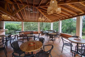 An outdoor patio area with wooden tables and chairs, large windows, and a view of greenery. - Tres Lagos Hotel (San Jerónimo)