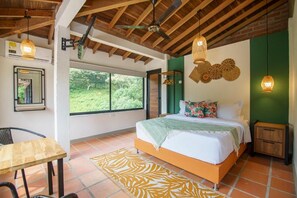 Un dormitorio con cama, una mesa, una silla, un ventilador de techo y una ventana con vista a la vegetación.