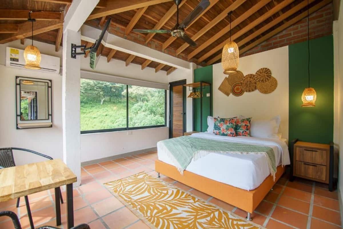 A bedroom with a bed, a table, a chair, a ceiling fan, and a window with a view of greenery.