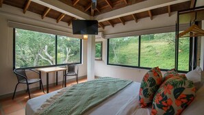 A bedroom with a bed, a desk, and two chairs. There are large windows providing a view of the outdoors. - Tres Lagos Hotel (San Jerónimo)