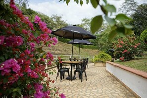 Une terrasse extérieure avec une table et des chaises, abritée par un parasol Club Colombia, et entourée d’une végétation luxuriante et de plantes en fleurs.