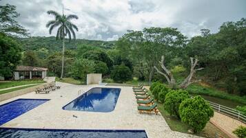 A swimming pool with lounge chairs, surrounded by greenery and a hill in the background.