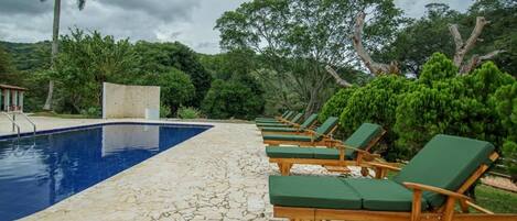 Uma área de piscina com piscina azul límpida, espreguiçadeiras de madeira e um deck com ladrilhos de pedra.