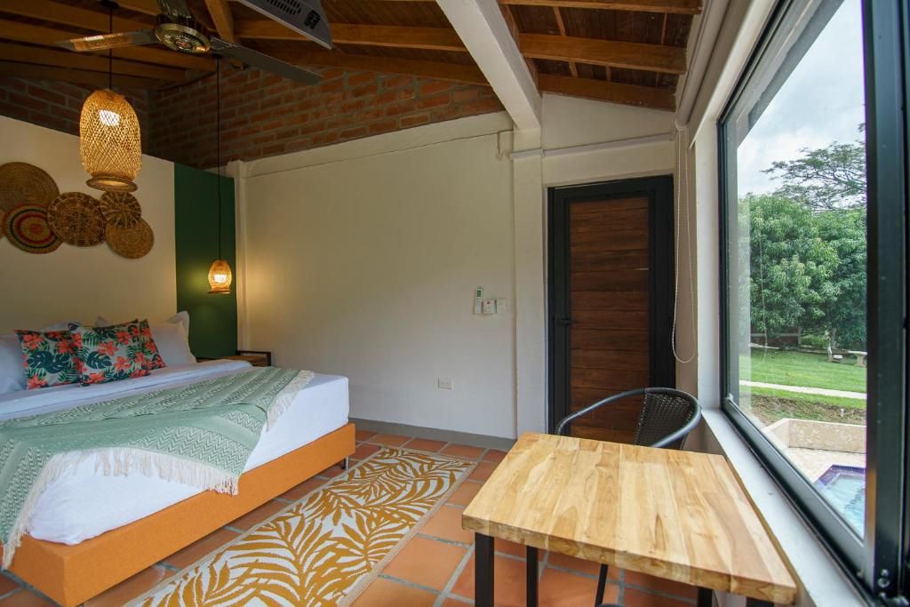 Un dormitorio con cama, una mesa de madera, una silla, una ventana con vistas a la vegetación y un ventilador de techo.
