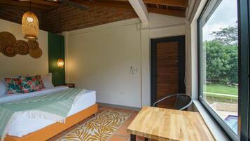 Un dormitorio con cama, una mesa de madera, una silla, una ventana con vistas a la vegetación y un ventilador de techo.