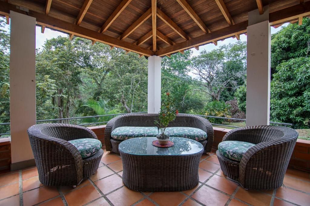 Un área de estar al aire libre techada, con mobiliario de mimbre y una mesa central, rodeada de exuberante vegetación.