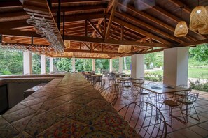Un espacio de comedor al aire libre cubierto con piso de baldosas, sillas de metal y lámparas colgantes.