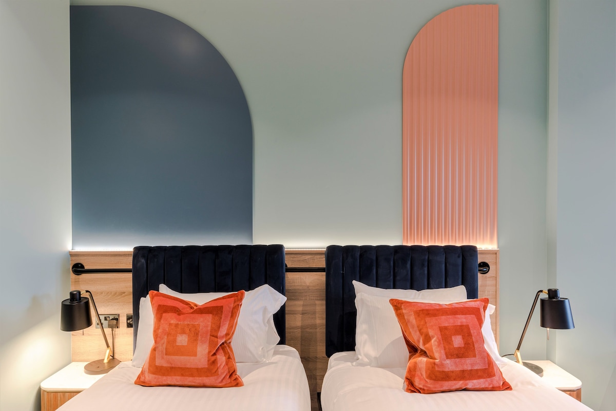 Une chambre d’hôtel avec deux lits, chacun doté d’une tête de lit sombre, de draps blancs et d’oreillers orange. On y trouve des tables de chevet avec des lampes, et une grande décoration murale abstraite au-dessus des lits.