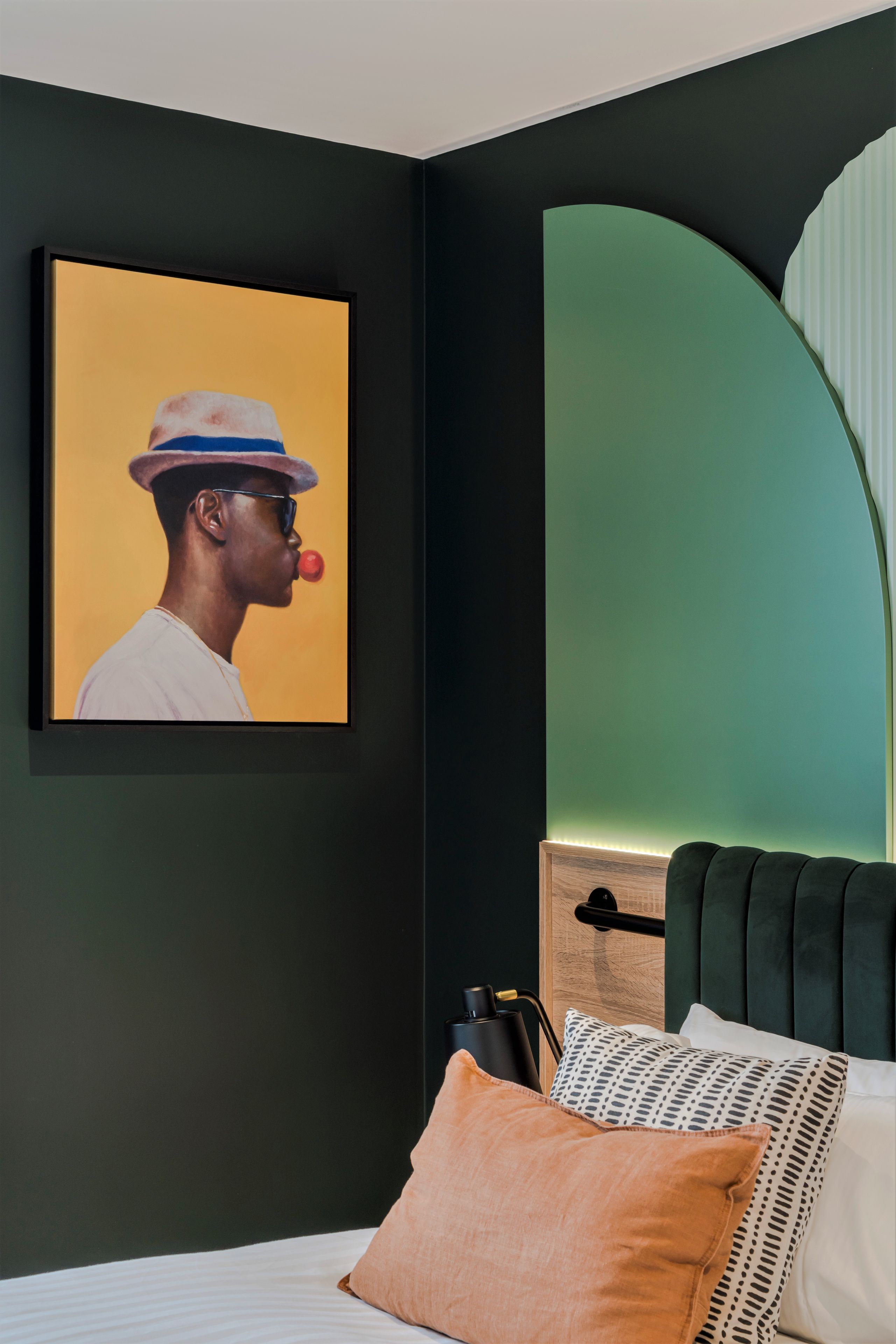 A bedroom with a dark green wall, a framed picture, a bed with a green upholstered headboard, and a white pillow.