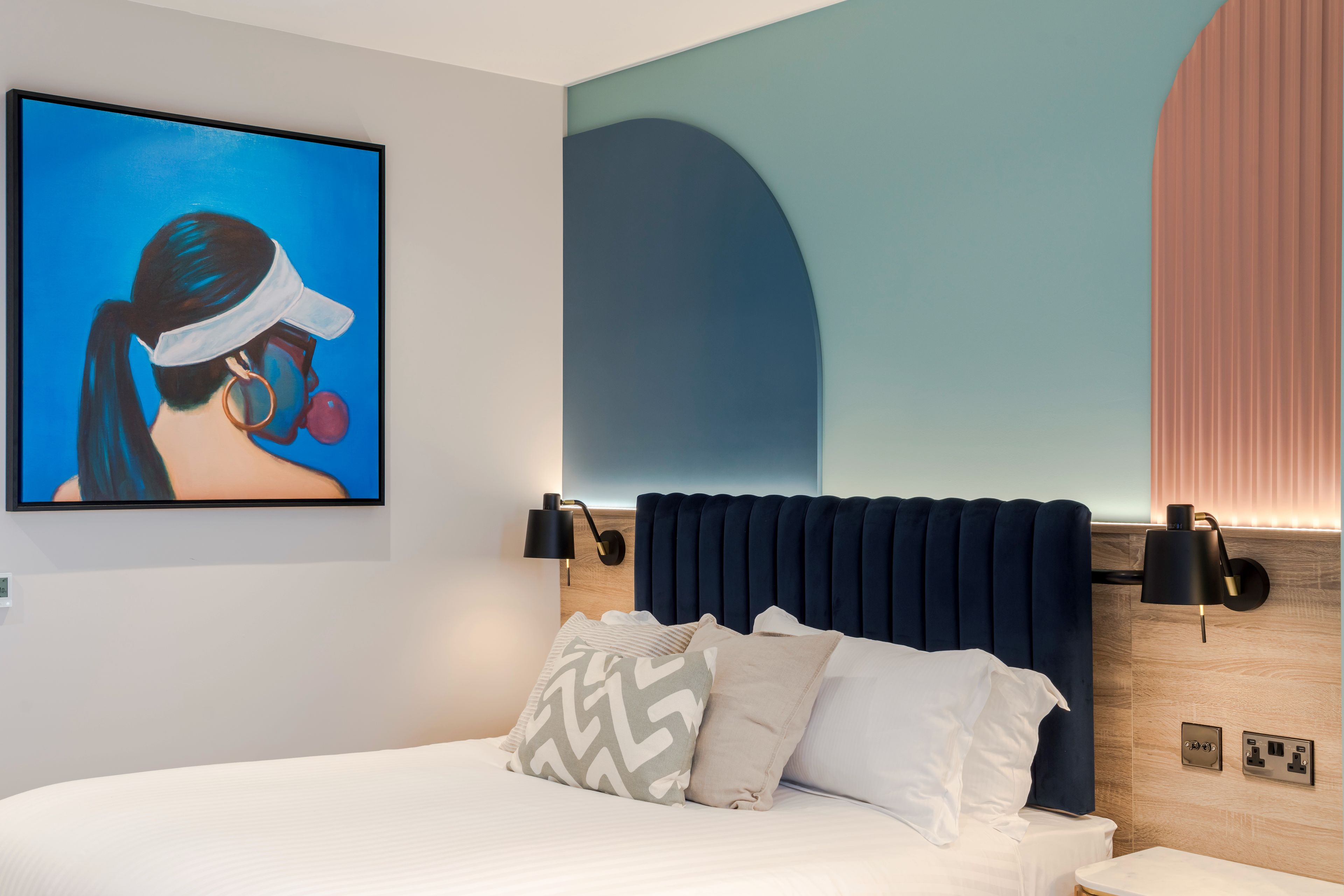 A modern hotel room with a large bed, a headboard with built-in lighting, a wall-mounted artwork, and a bedside table.