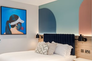 Una habitación de hotel moderna con una cama grande, cabecera con iluminación integrada, una obra de arte colgada en la pared y una mesita de noche.