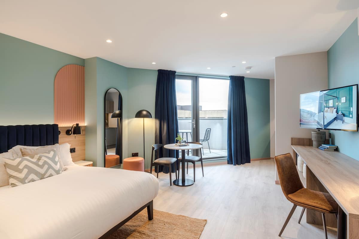 Une chambre d’hôtel moderne équipée d’un lit, d’un bureau, d’une chaise, d’une table ronde et d’un balcon avec des rideaux.