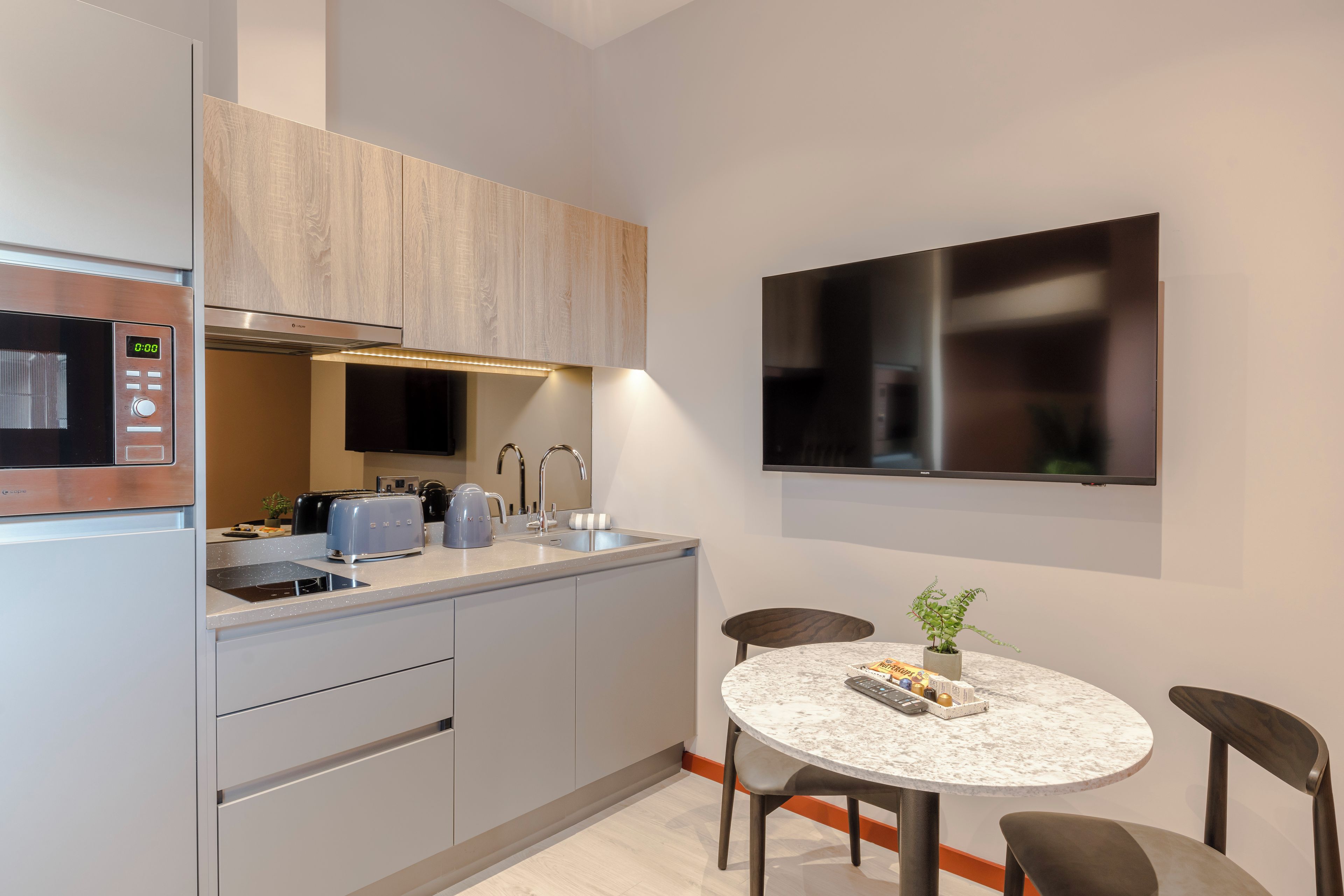 Una cocina moderna con microondas integrado, tostadora y lavamanos. Hay una mesa redonda con sillas y una pequeña planta en maceta sobre la encimera.