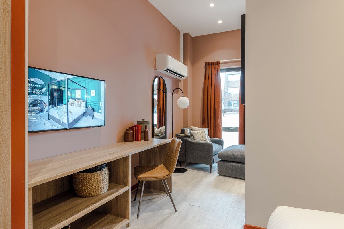 Un salon moderne avec un bureau en bois, un fauteuil confortable, un canapé gris et un grand téléviseur à écran plat fixé au mur.