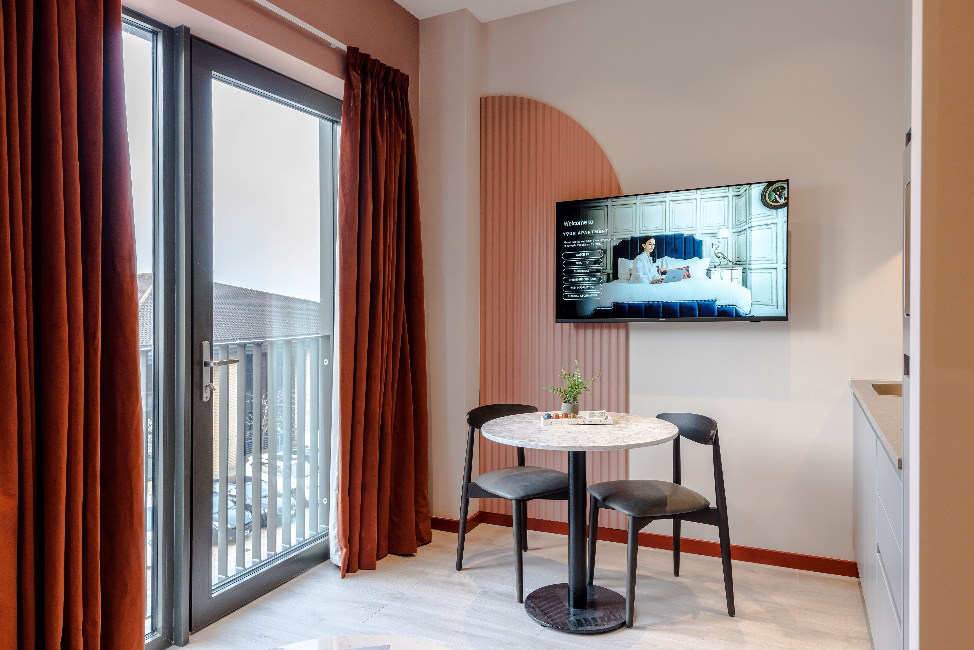 A modern room with a sliding glass door, a round table with two chairs, and a flat-screen TV mounted on the wall displaying a welcome message.