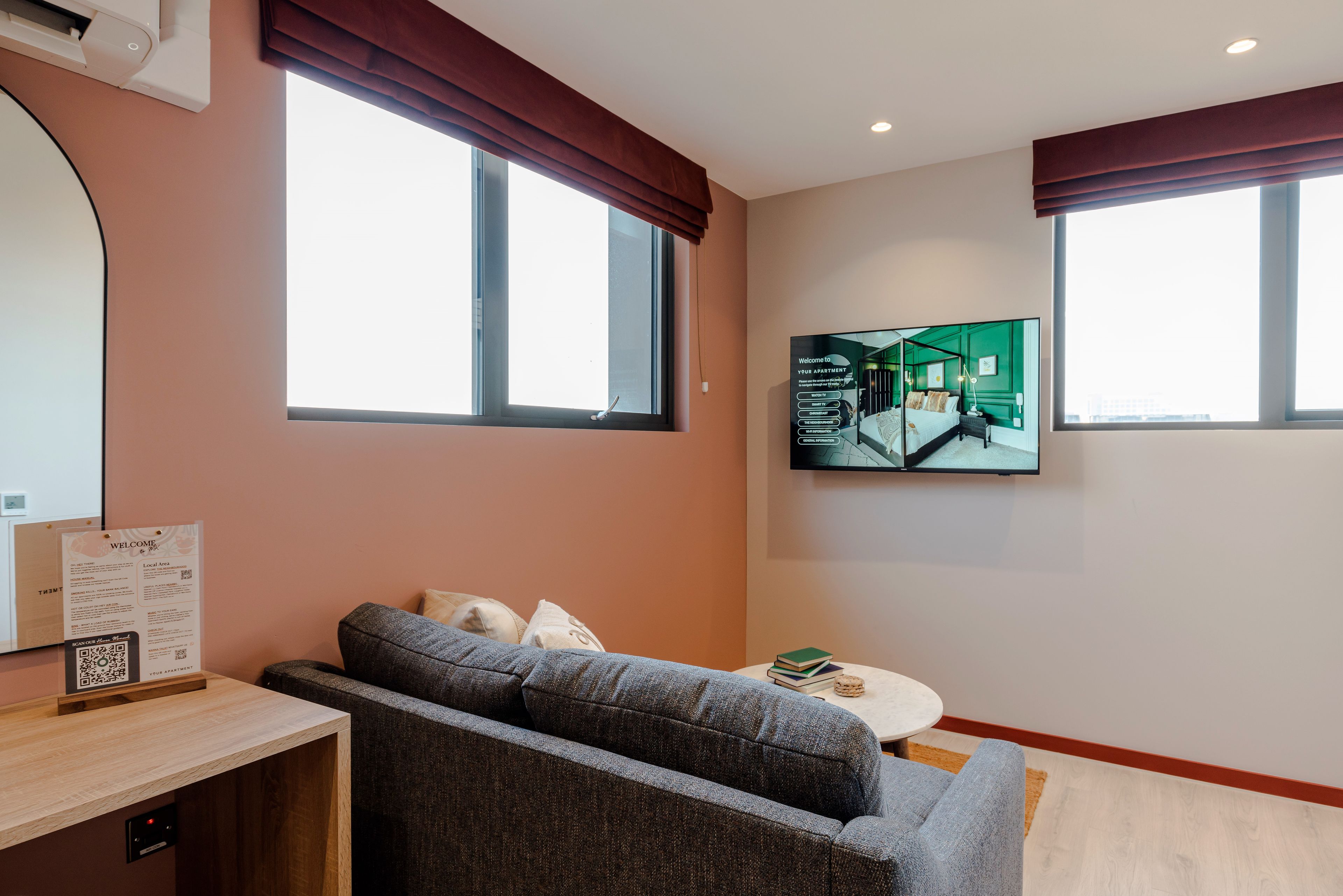 A modern living room with a grey sofa, a small round table with books, a mounted TV, and a large window with a view of the building's interior.