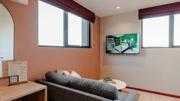 A modern living room with a grey sofa, a small round table with books, a mounted TV, and a large window with a view of the building's interior.