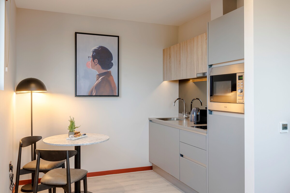Une cuisine compacte équipée d’un four à micro-ondes, d’un évier et de placards. On y trouve une table ronde avec des chaises et une petite plante. Un tableau encadré est accroché au mur.