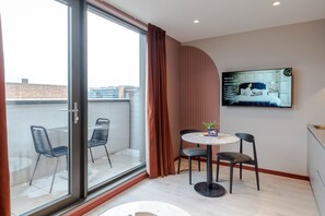 Une chambre moderne dotée d’une porte-fenêtre coulissante donnant sur un balcon, d’une table de salle à manger ronde avec des chaises et d’un téléviseur à écran plat fixé au mur.