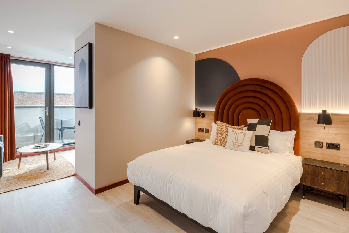 Une chambre moderne avec un grand lit, une tête de lit en bois et une vue sur la ville grâce à de grandes fenêtres.