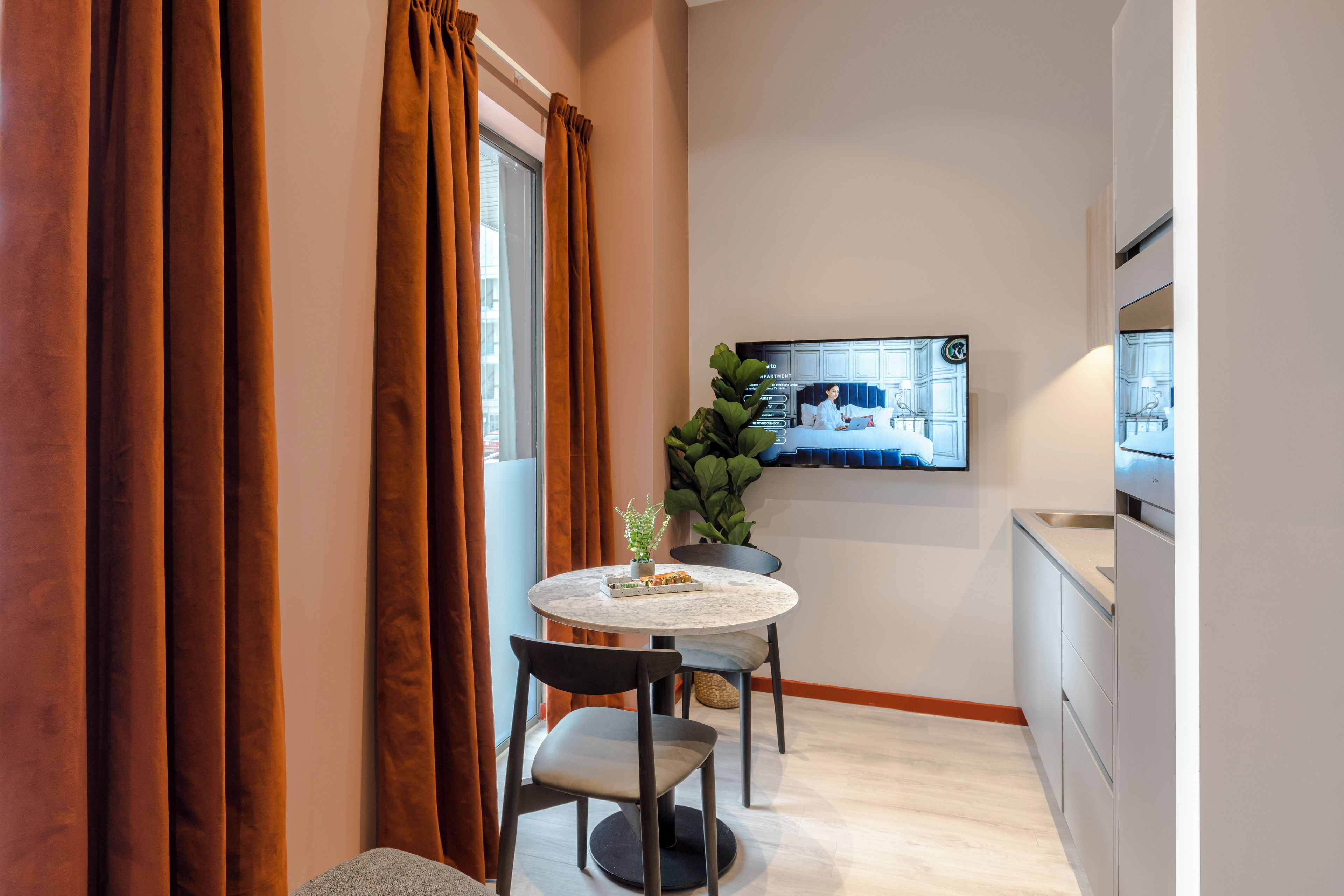 A compact kitchen with a dining area, featuring a round table, two chairs, a potted plant, and a flat-screen TV mounted on the wall.